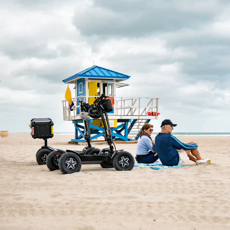 Lifestyle view of the MIA Four X2 scooter parked on the beach near a couple sitting on a blanket